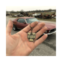 V8 Auto Schlüsselanhänger in der Hand vor Muscle‑Car Hand präsentiert einen V8 Eight Schlüsselanhänger aus Messing vor einem US‑Car‑Schrottplatz, ideal für Gearheads, Hot‑Rod‑ und V8‑Fans.