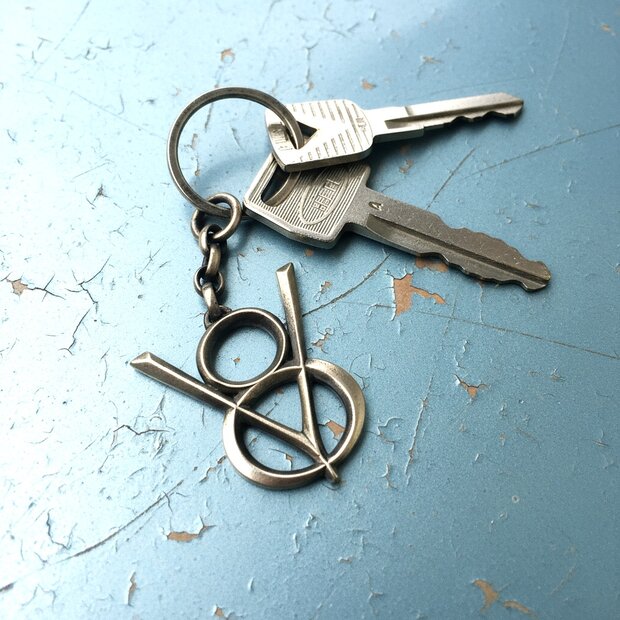 V8 Schlüsselanhänger – Sterling, Vintage Look Silberner V8-Anhänger am Schlüsselbund mit klassischem Retro Design, inszeniert auf blauer Haube mit Lackrissen.