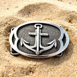 Ovale, handgefertigte Gürtelschnalle aus massivem Silber mit detailreichem Anker-Motiv auf strukturiertem, dunkel patiniertem Hintergrund. Liegt im Sand.